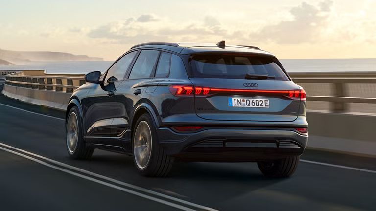 Schräge Heckaufnahme des Q6 SUV e-tron, welche über eine kurvige Straße am Meer fährt. Im Hintergrund sind Berge zu sehen.