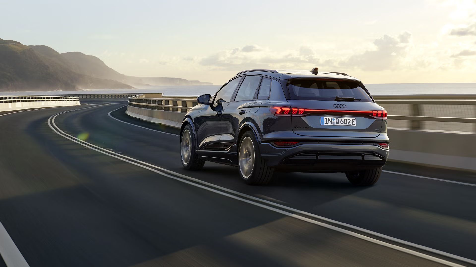 Schräge Heckaufnahme des Q6 SUV e-tron, welche über eine kurvige Straße am Meer fährt. Im Hintergrund sind Berge zu sehen.