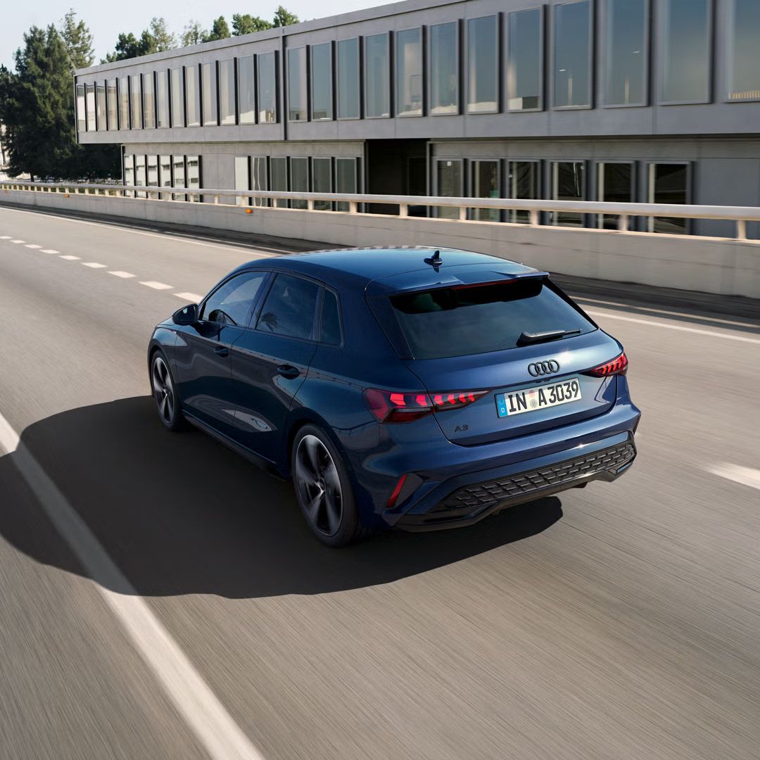 Der Audi A3 Sportback schräg von hinten auf einer Brücke, die einfallende Sonne wirft auf der linken Fahrzeugseite einen großen Schatten. Im Hintergrund ein Bürogebäude.