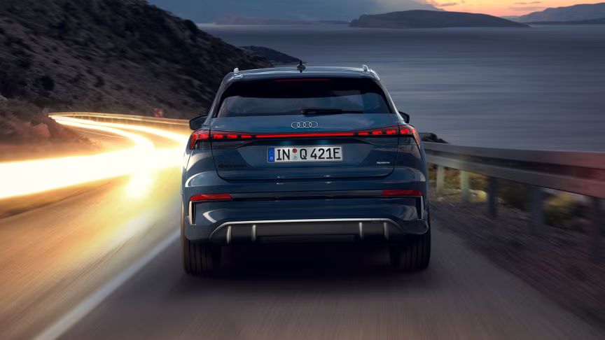 Der Q4 SUV e-tron von hinten bei Nacht auf einer abschüssigen Gebirgstraße, zur rechten das Meer. Auf der Gegenfahrbahn sind Lichtschleier entgegenkommender Fahrzeuge zu sehen. 