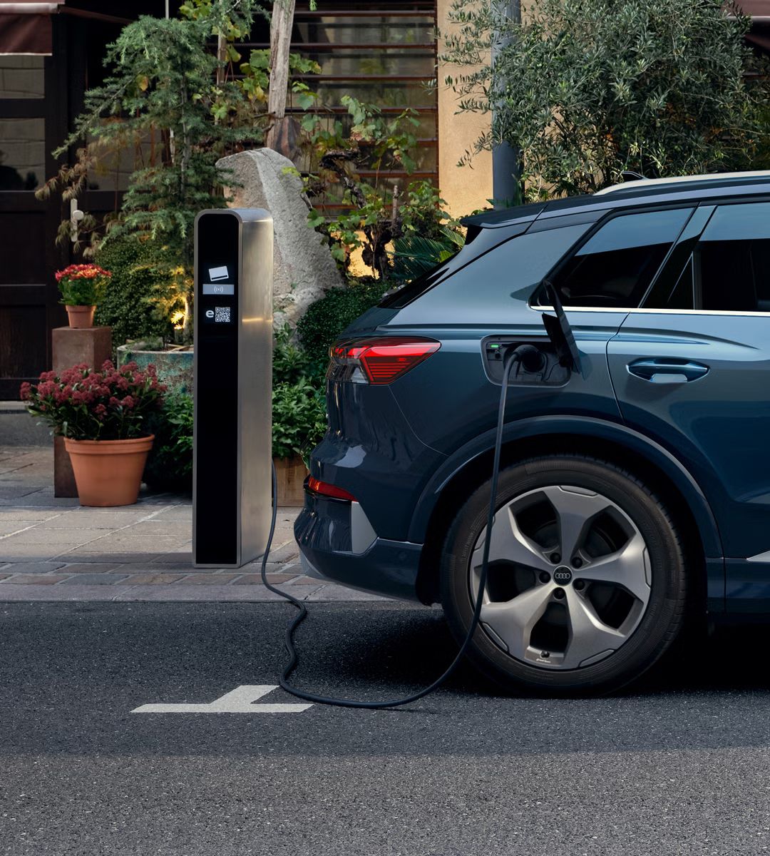 Nahaufnahme des rechten Hecks des Q4 SUV e-tron, parkend vor einem Gebäude, Pflanztöpfe sind vor dem Gebäude zu sehen. Das Fahrzeug ist an eine öffentliche Ladesäule angeschlossen. 